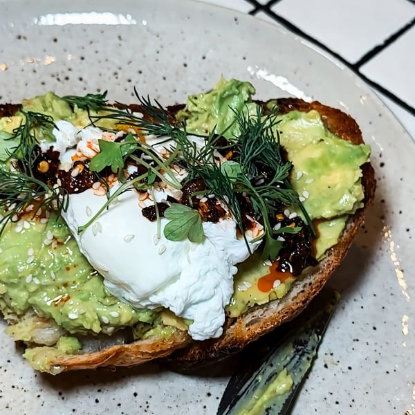 Avocado toast with a poached egg, chili oil, sesame seeds, and fresh herbs on a white plate.