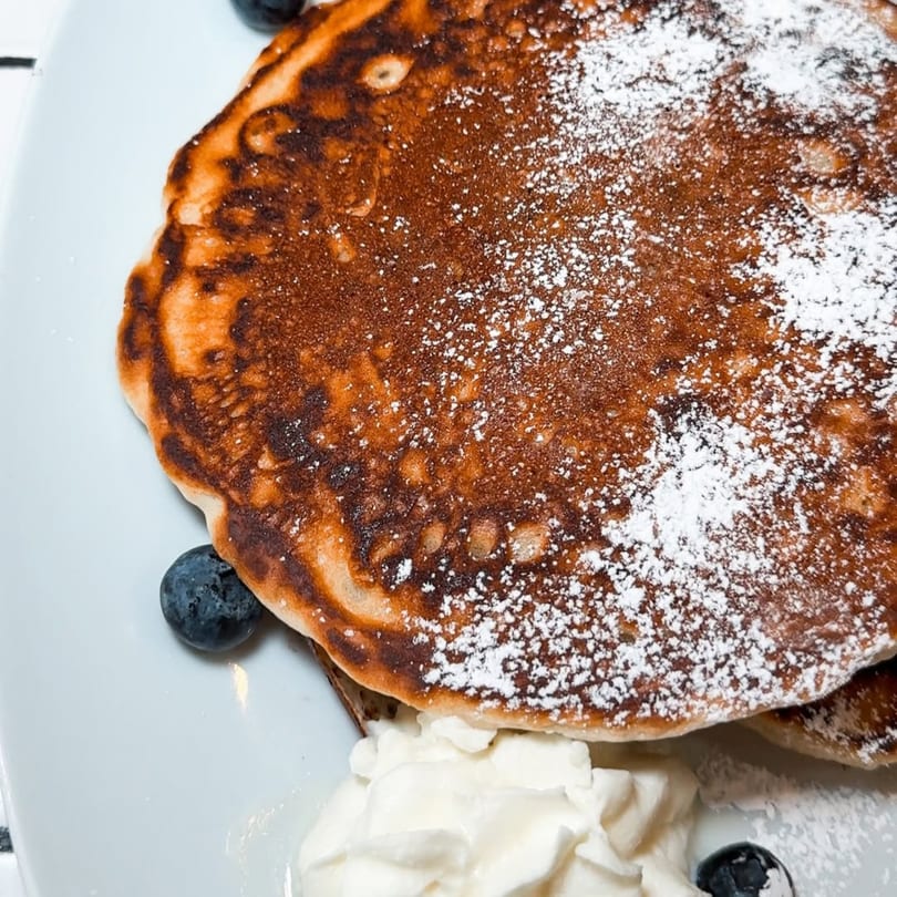 Fluffy pancakes topped with powdered sugar, fresh blueberries, and whipped cream on a white plate.