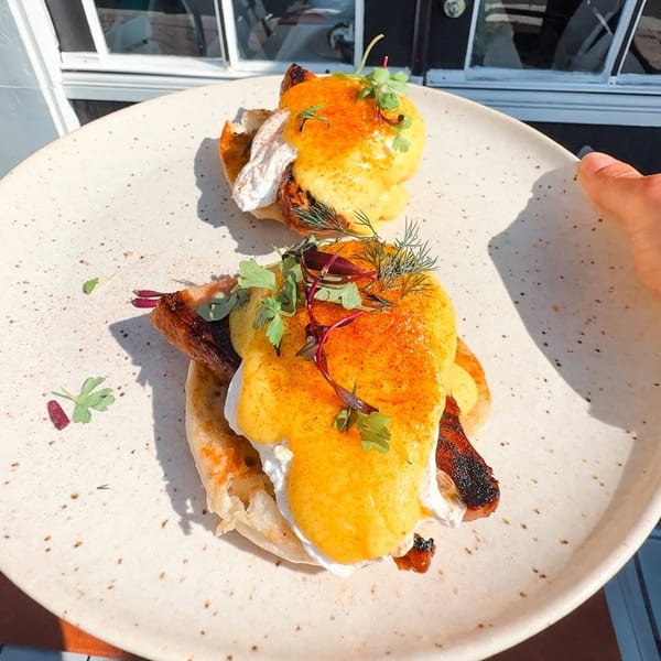 Two servings of eggs Benedict with bacon, hollandaise sauce, and herbs on a speckled plate.